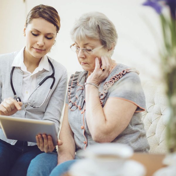 Latin nurse with a stethoscope around her neck is consulting with a senior lady on a sofa. As she speaks, she is smiling and sharing the examination results to her patient.